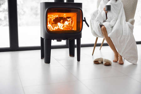 Woman sitting on chair by the burning fireplace at home. Concept of relaxing and leisure time at home in winter time. Cropped view without face focused on legs and fireplaceの写真素材