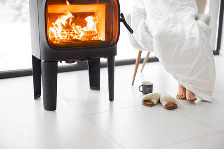 Woman sitting on chair by the burning fireplace at home. Concept of relaxing and leisure time at home in winter time. Cropped view without face focused on legs and fireplaceの写真素材