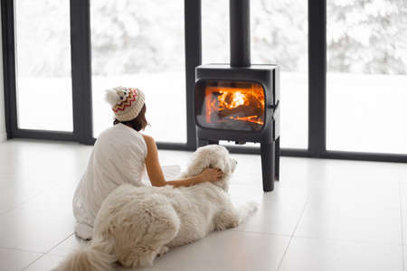 Woman sitting with her dog by the fireplace at modern house on nature during winter time. Concept of winter mood and comfort at home. Girl in hat covered with blanletの写真素材
