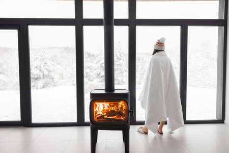 Woman enjoys winter time at home with burning fireplace and panoramic window, looking outside on snowy landscape. Concept of winter mood and comfort at home. Idea of winter vacation in the mountainsの写真素材