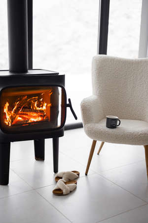 Cozy living space by the burning fireplace with chair, cup and slippers on background of snowy landscape. Idea of home comfort and winter moodの写真素材