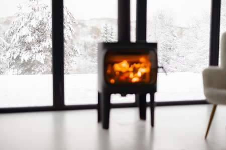 Cozy living space by the burning fireplace near window with beautiful snowy landscape behind. Idea of home comfort and winter mood. Rest in modern cabins on natureの写真素材