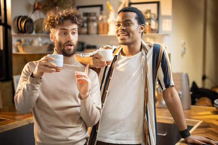 Two guys of different ethnicity having warm conversation while drinking coffee on kitchen at home. Concept of close male friendship or relationship as gay couple. Caucasian and hispanic man togetherの写真素材