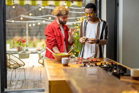 Two brightly dressed stylish guys having fun while making salad together on background of backyard. Concept of gay couples and everyday life at home. Caucasian and hispanic man cooking healthy foodの写真素材