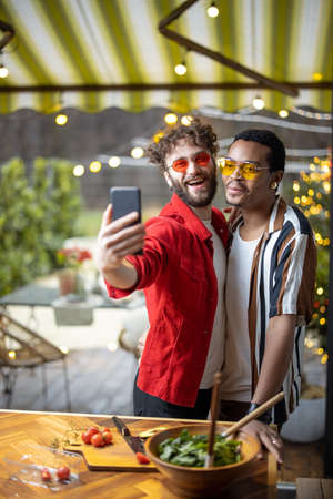 Two brightly dressed stylish guys taking selfie on phone, while having fun together at home. Concept of multiracial diverse gay couple and style. Caucasian and hispanic man having close relationshipの写真素材