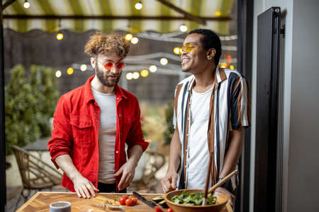 Two brightly dressed stylish guys having fun while making salad together indoors on background of backyard. Concept of gay couples. Caucasian and hispanic man having close conversationの写真素材