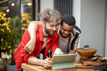 Two brightly dressed stylish guys watching a movie on digital tablet, hugging together at cozy modern home. Concept of gay couples. Caucasian and hispanic man having fun togetherの写真素材