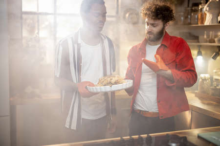 Portrait of two lovely multiracial guys standing together with cooked turkey on kitchen with a steam on background from the oven. Concept of gay relations everyday life together at homeの写真素材