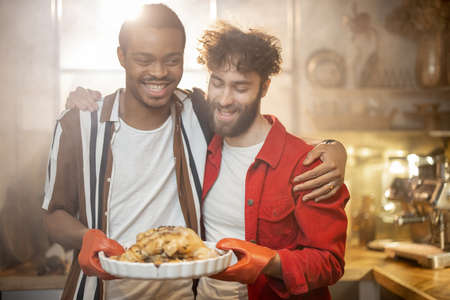 Portrait of two lovely multiracial guys standing together with cooked turkey on kitchen with a steam on background from the oven. Concept of gay relations everyday life together at homeの写真素材