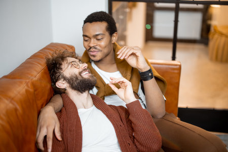 Gay couple with different nationality having close conversation while sitting on a couch at home. Concept of homosexual relations and lifestyle at home. Caucasian and hispanic man together indoorsの写真素材