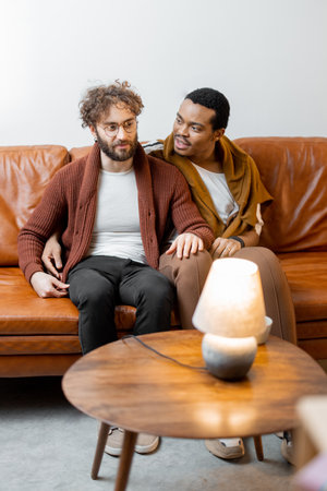 Gay couple with different nationality having close conversation while sitting on a couch at home. Concept of homosexual relations and lifestyle at home. Caucasian and hispanic man together indoorsの写真素材