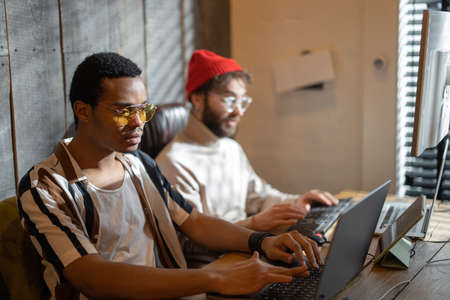 Two men with different nationality working on computers, sitting together at cozy home office. Concept of freelance and remote work. Stylish male hipsters programming togetherの写真素材