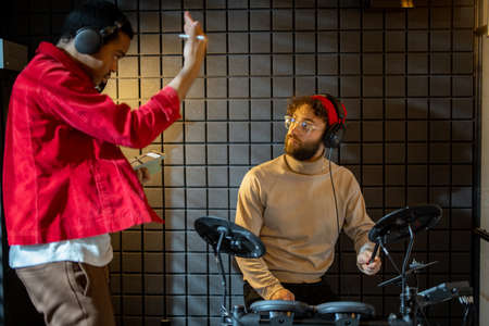 Two stylish men playing electric drums, composing electronic music at small recording studio. Small band creating or practicing music in a home studio. Caucasian and hispanic man playing togetherの写真素材