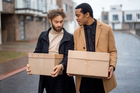Stylish gay couple walking together with parcels on some residential street on rainy weather. Caucasian and hispanic man wearing coats. Concept of homosexual lifestyle and deliveryの写真素材