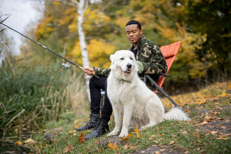 Latin man sitting in chair and fishing near Maremmano-Abruzzese Sheepdog on river or lake coast. Concept of leisure, hobby, vacation and weekend. Man resting in nature at autumn dayの写真素材