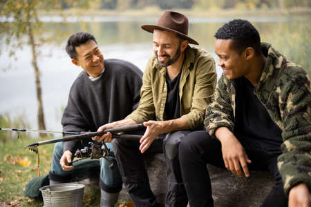 Multiracial male friends resting and talking in nature. Men after fishing on river or lake coast. Concept of leisure and weekend in nature. Idea of friendship and spending time together with dogの写真素材