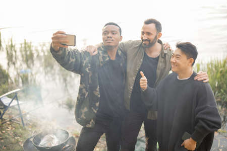 Smiling multiracial male friends taking selfie on smartphone during resting in nature. Men fishing and cooking Ukha soup on river coast at autumn. Leisure, weekend and vacation. Idea of friendshipの写真素材