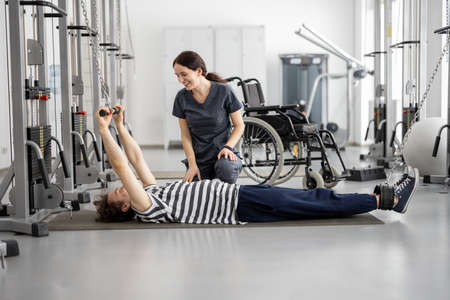 Rehabilitation specialist helps a guy to do exercise on decompression simulator for recovery from injury. Wheelchair on background. Concept of physical therapy for people with disabilitiesの写真素材