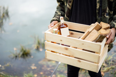 Man holding wooden box with firewoods and bottles full of alcoholic drink. Concept of leisure in nature with alcoholic beveragesの写真素材