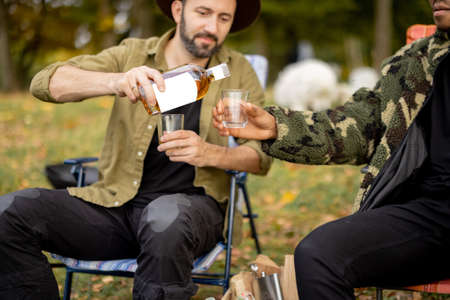 Two guys pouring alcohol in glasses, while fishing together on the lake coast. Caucasian and latin man resting on nature. Concept of male friendship and alcoholic drinksの写真素材