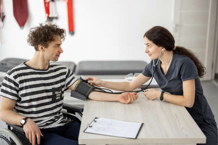 Medical worker measures the pressure of a young man in a wheelchair before training in the rehabilitation center. Concept of medical care and support for people with injuriesの写真素材