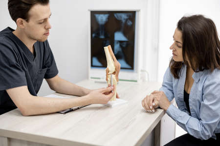 Physical therapist showing model of knee joint to a young woman patient during consultation at medical office. Concept of orthopedic appointment and treatmentの写真素材