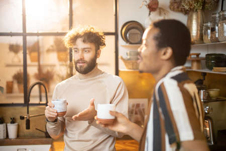 Two guys of different ethnicity having warm conversation while drinking coffee on kitchen at home. Concept of close male friendship or relationship as gay couple. Caucasian and hispanic man togetherの写真素材