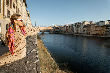 Young woman enjoys beautiful view on famous Old bridge in Florence, sitting on the riverside at sunset. Female traveler visiting italian landmarks. Stylish woman wearing dress and colorful shawlの写真素材