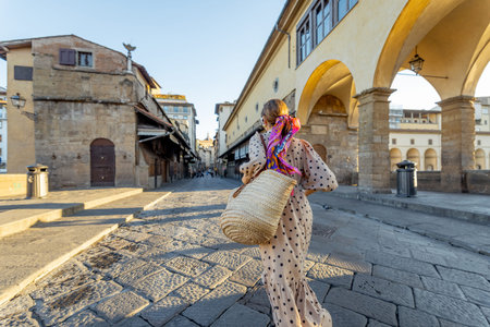 Woman walking on famous old bridge, called Ponte Vecchio, in Florence. Concept of traveling italian landmarks. Stylish woman with colorful shawl and sunglasses. Wide view on sunriseの写真素材
