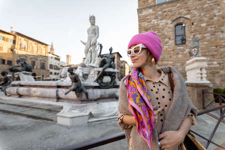 Young woman traveling famous italian landmarks in Florence city. Enjoying beautiful architecture and Neptuine fountain on Signoria square. Woman dressed in Italian style with colorful scarf and hatの写真素材
