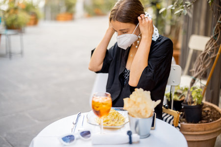 Young woman takes off face mask going to sit at restaurant table outdoors on cozy street in Italy. Concept of social rules during pandemic and italian lifestyleの写真素材