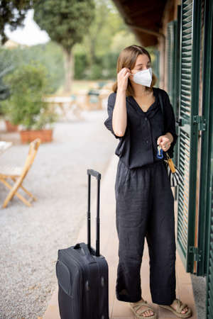 Woman in medical mask with baggage and keys go in hotel room. Concept of weekend, rest, tourism and vacation. Idea of health protection during pandemic. Young caucasian girl looking awayの写真素材