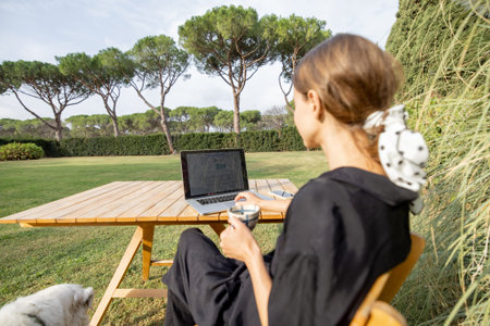 Young caucasian woman watching something on laptop computer and drinking tea while sitting at wooden table on green mediterranean lawn. Concept of weekend, rest and remote workの写真素材