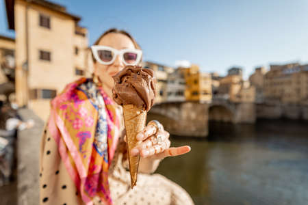 Young woman eating italian ice cream while sitting on the riverside with beautiful view on famous Old bridge in Florence at sunset. Concept of italian gastronomy and landmarksの写真素材