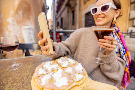 Woman having fun while eating pizza and drinking cocktail at restaurant on a street in Rome. Concept of Italian gastronomy and travel. Stylish woman with sunglasses and colorful hair shawlの写真素材