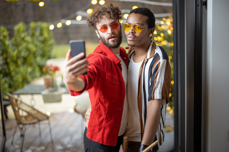 Two brightly dressed stylish guys taking selfie on phone, while having fun together at home. Concept of multiracial diverse gay couple and style. Caucasian and hispanic man having close relationshipの写真素材