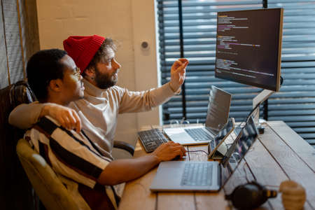 Two men with different nationality programming on computers, sitting at cozy home office. Concept of freelance and remote work. Iea of gay couples working together. Stylish male hipsters programming togetherの写真素材