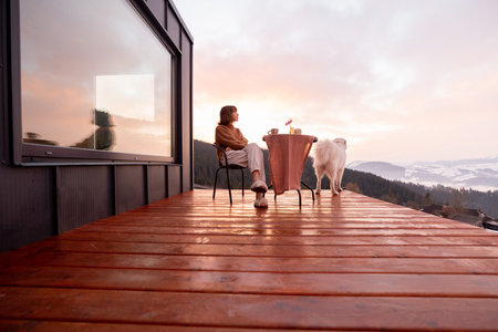 Woman with her dog resting on terrace of tiny house in the mountains, enjoying beautiful landscape during sunrise. Concept of small modern cabins for rest and escape to natureの写真素材