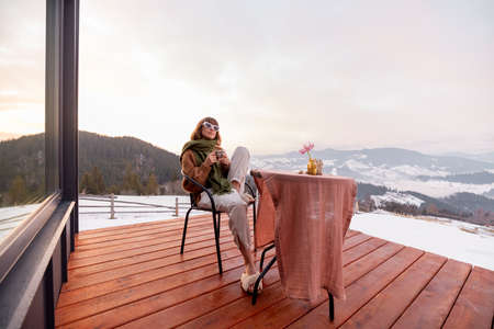 Woman sitting on terrace with great view on the mountains in winter, enjoying drink and beautiful landscape during sunrise. Concept of rest and escape to natureの写真素材