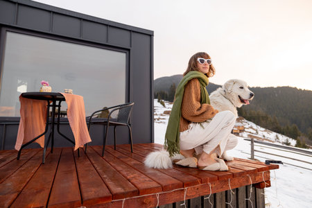 Woman with her dog resting on terrace of tiny house in the mountains on winter time. Concept of...