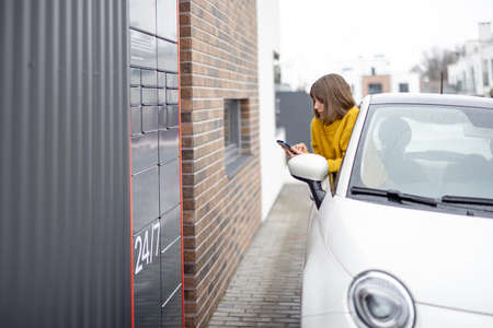 Woman opens post terminal cell contactlessly using smart phone right out of the car window. Concept of fast delivery and receiving goods on the goの写真素材