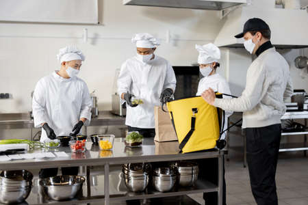 Courier waiting for an order for delivery in the kitchen with chefs preparing takeaway foodの素材 [FY310183038469]