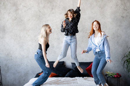 Adult girlfriends having fun, jumping together on bed at homeの写真素材