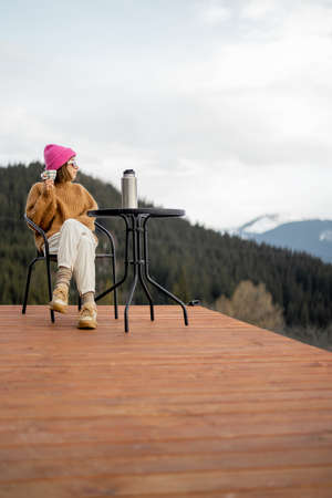 Woman enjoys great view on mountains, sitting on terrace and drinking hot drink. Concept of escape and recreation on natureの写真素材
