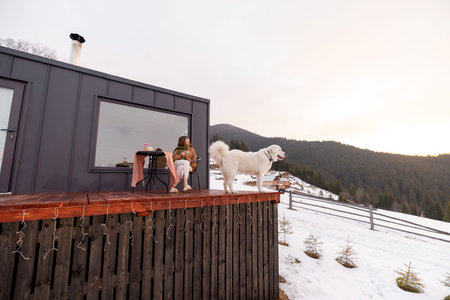Woman with her dog resting on terrace of tiny house in the mountains on winter time. Concept of...