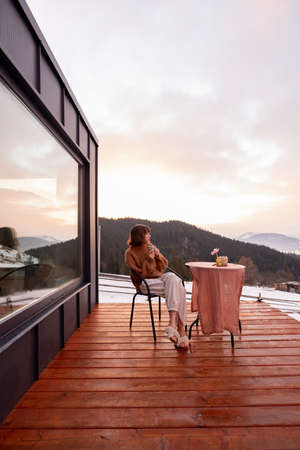 Woman having a breakfast while resting on terrace of tiny house in the mountains, enjoying beautiful landscape during sunrise. Concept of small modern cabins for rest and escape to natureの写真素材