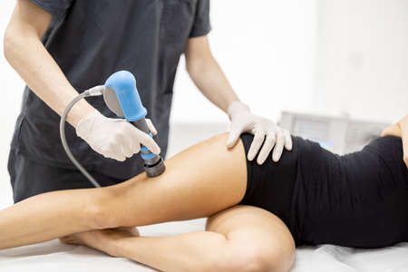 Doctor applies shock wave therapy with special medical equipment on womens thigh muscle at medical office. Concept of non-invasive technology for treating pain in musculoskeletal systemの写真素材