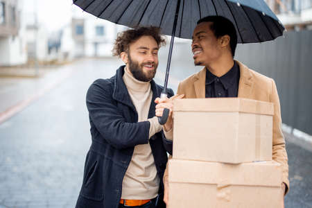 Stylish gay couple walking together with parcels on some residential street on rainy weather. Caucasian and hispanic man with umbrella wearing coats. Concept of homosexual lifestyle and deliveryの写真素材