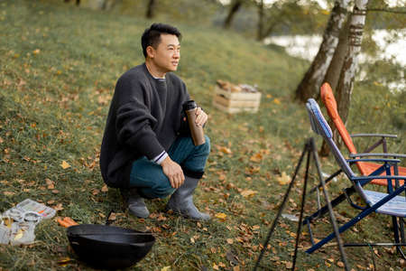 Asian man resting while cooking and fishing on lake shore. Concept of leisure, hobby and weekend in nature. Autumn dayの写真素材