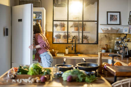 Woman looks into a fridge while cooking in the kitchen at home, healthy food on table top in front. Modern kitchen interior with a windowの写真素材